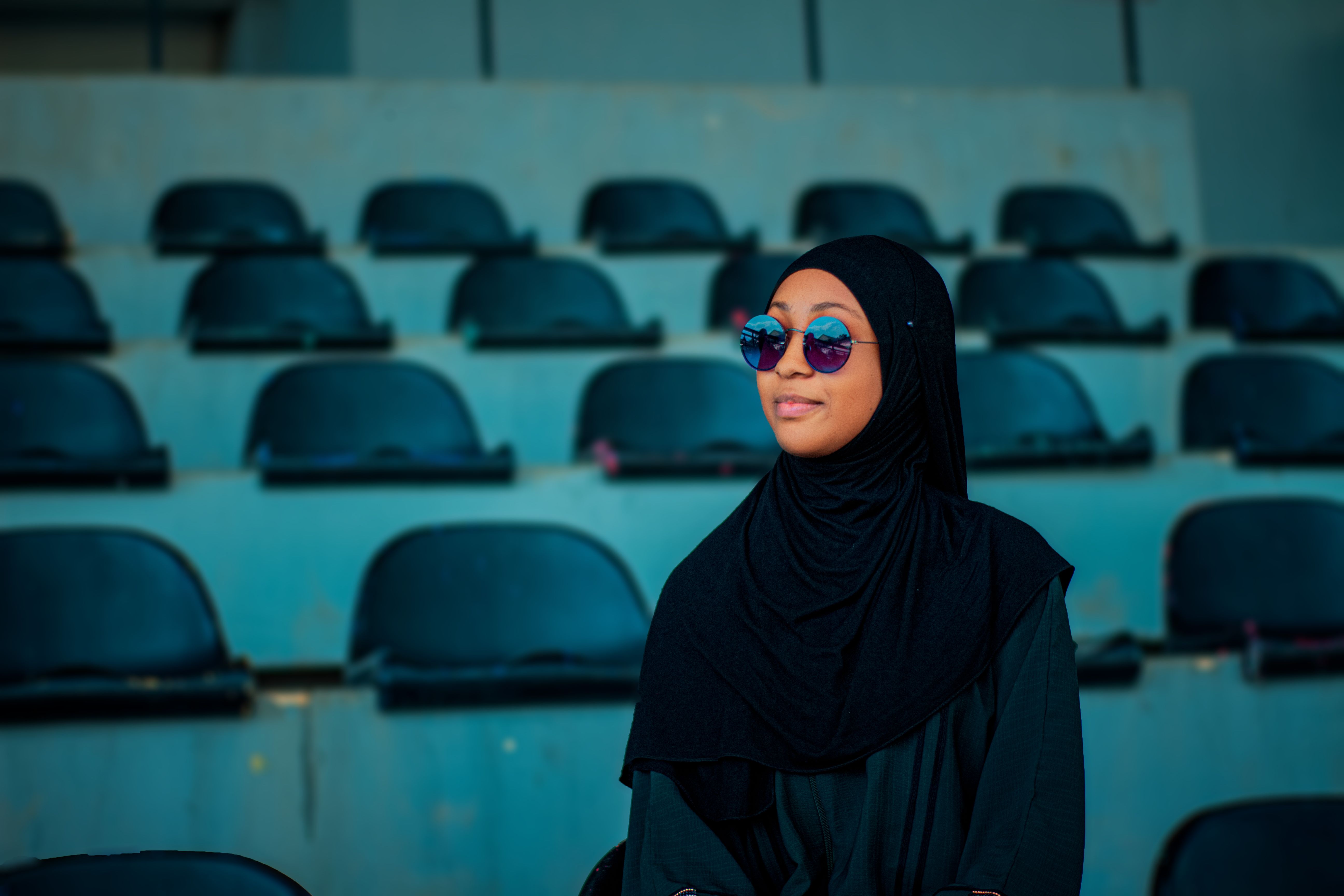 Stylish woman in black hijab and sunglasses sitting in empty stadium seats.