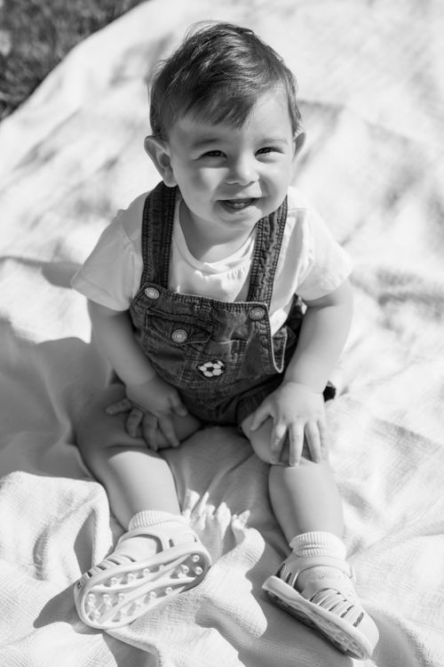 Free Black and white photo of a smiling baby sitting on a blanket outdoors, wearing overalls. Stock Photo