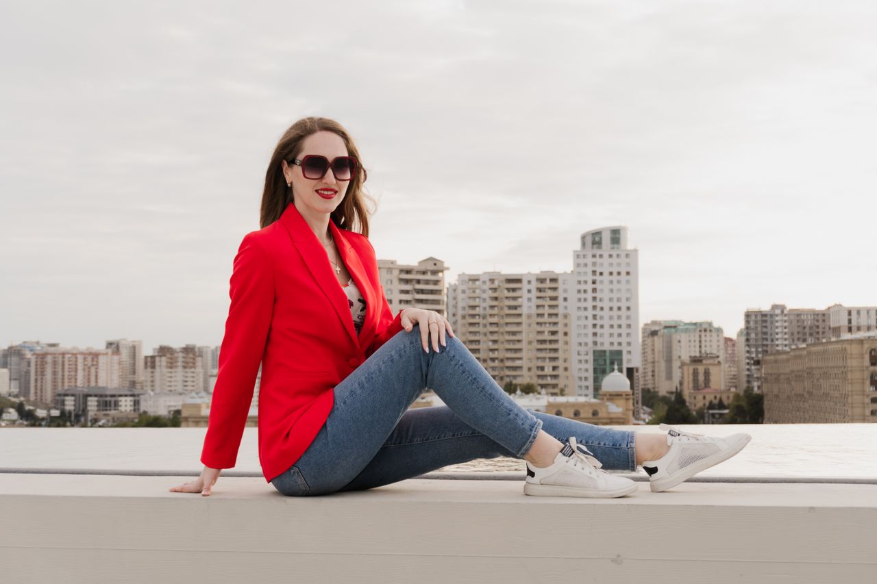 Woman in a red blazer, graphic tee, blue jeans, and white sneakers sitting on a rooftop ledge with a city skyline behind her.