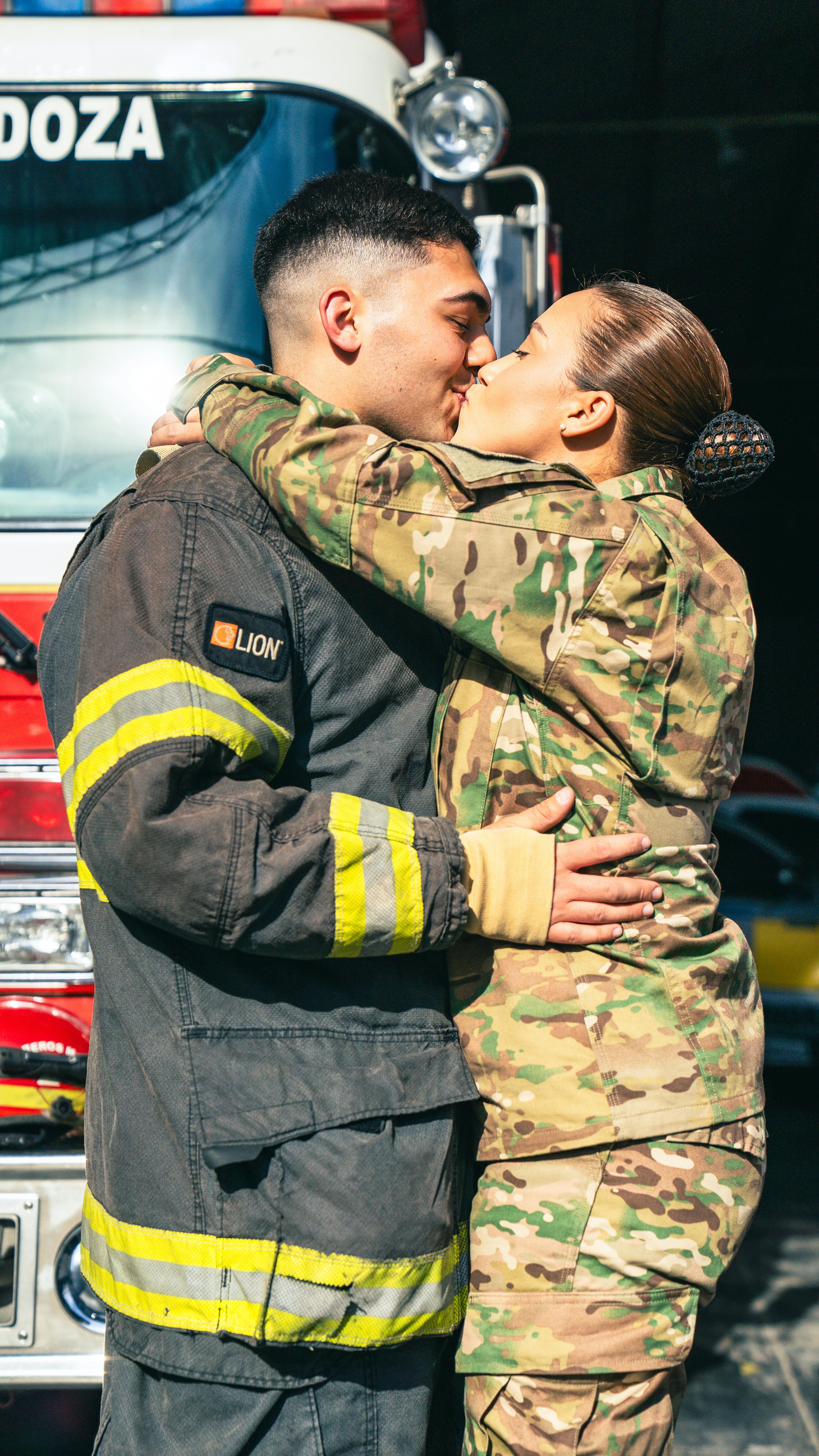 A firefighter and solider share a passionate kiss in front of a fire truck, symbolizing love and service.