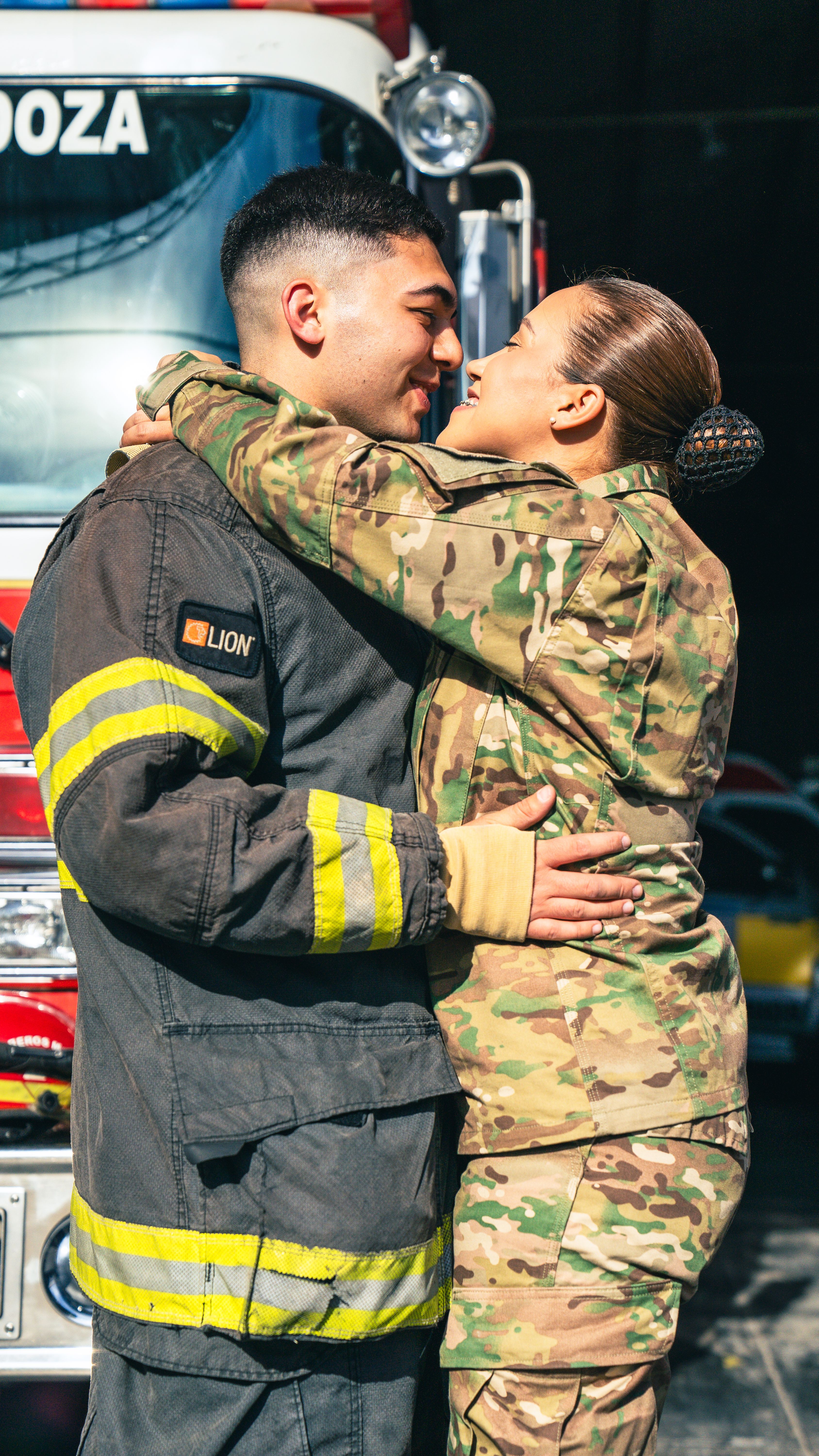 A firefighter and soldier embrace joyfully in front of a fire truck, symbolizing love and unity.