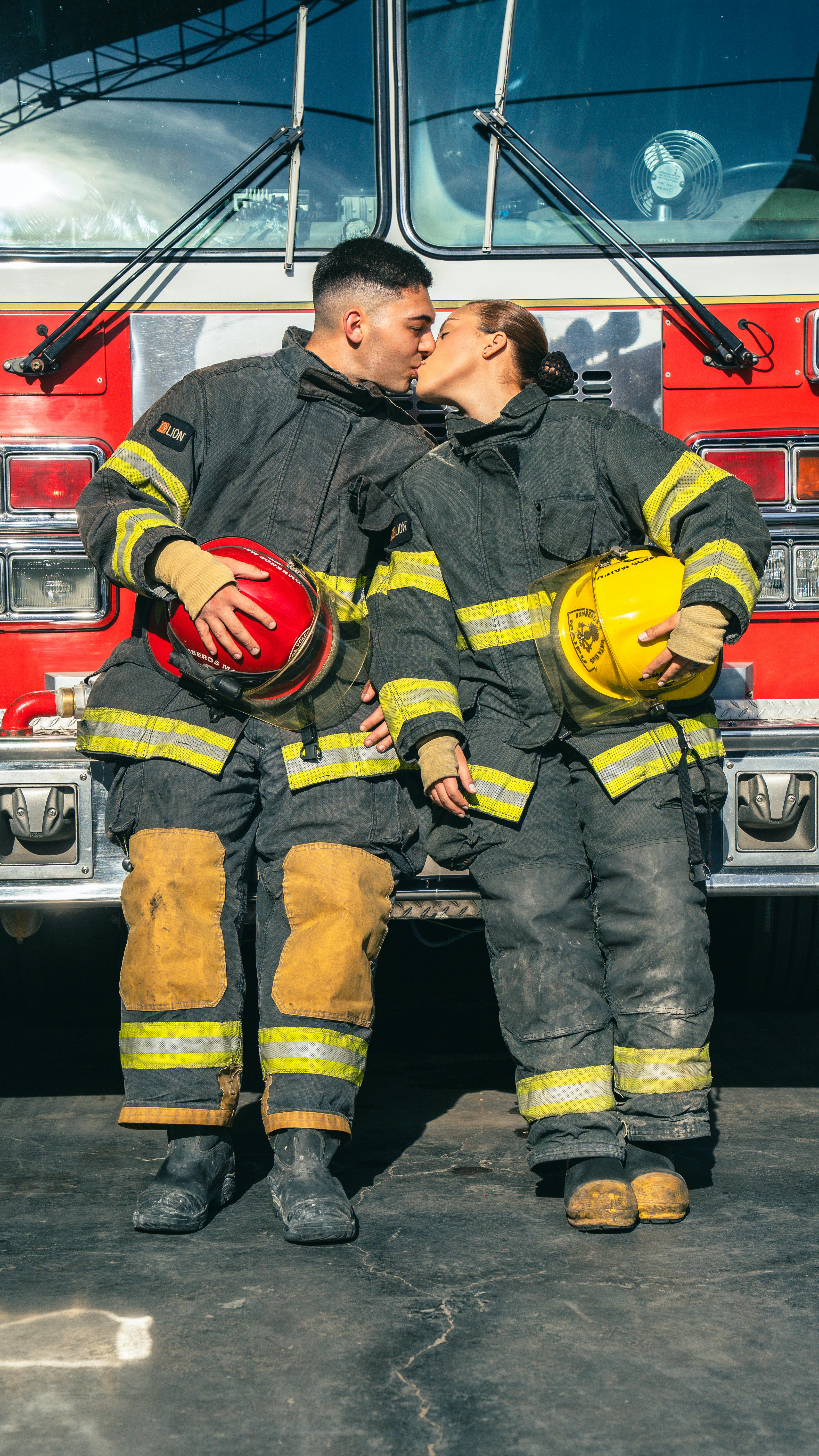 Two firefighters share a kiss in front of a fire truck, holding their helmets.