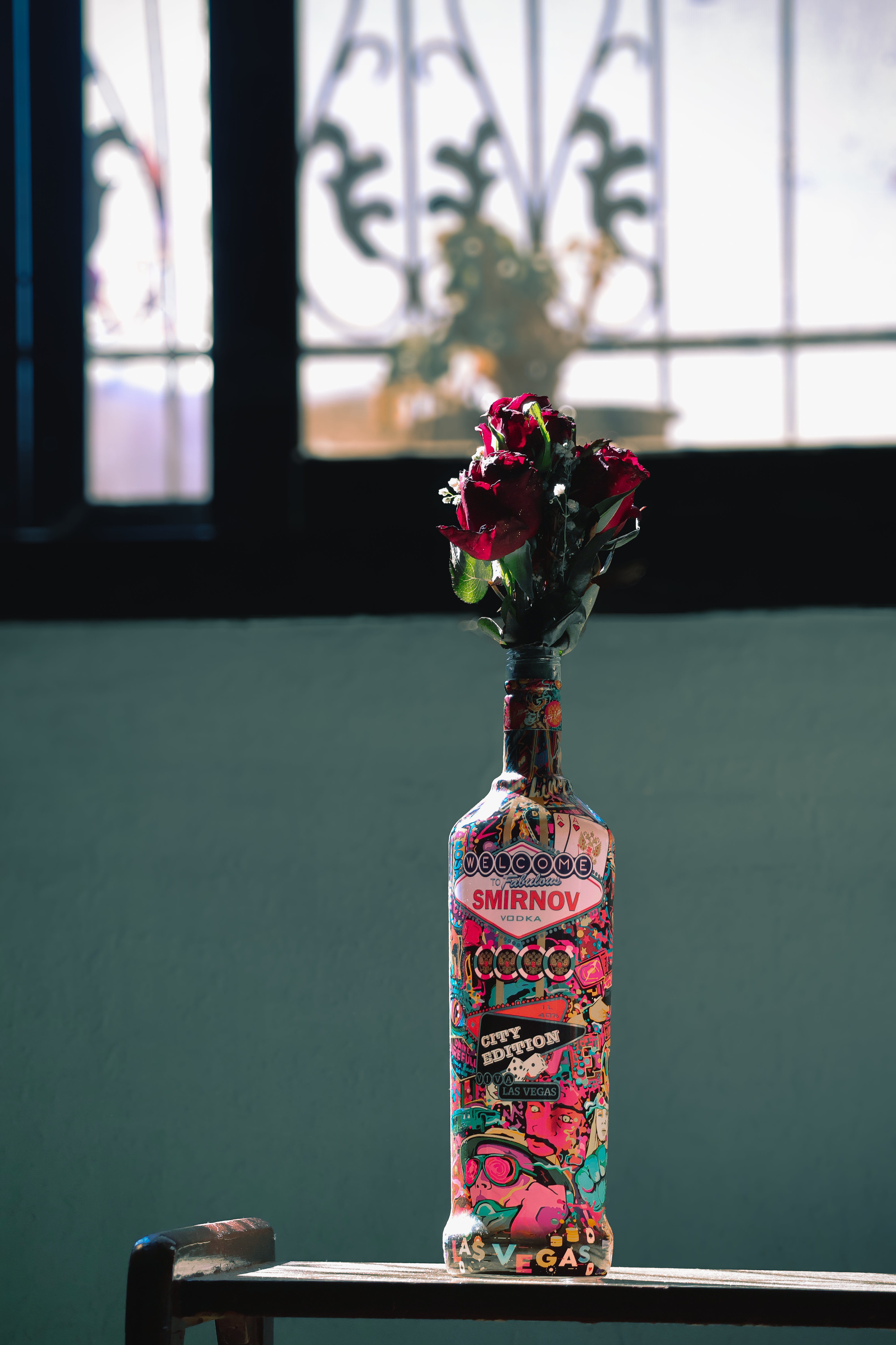 Artistic bottle vase with red roses on a table by a window indoors.