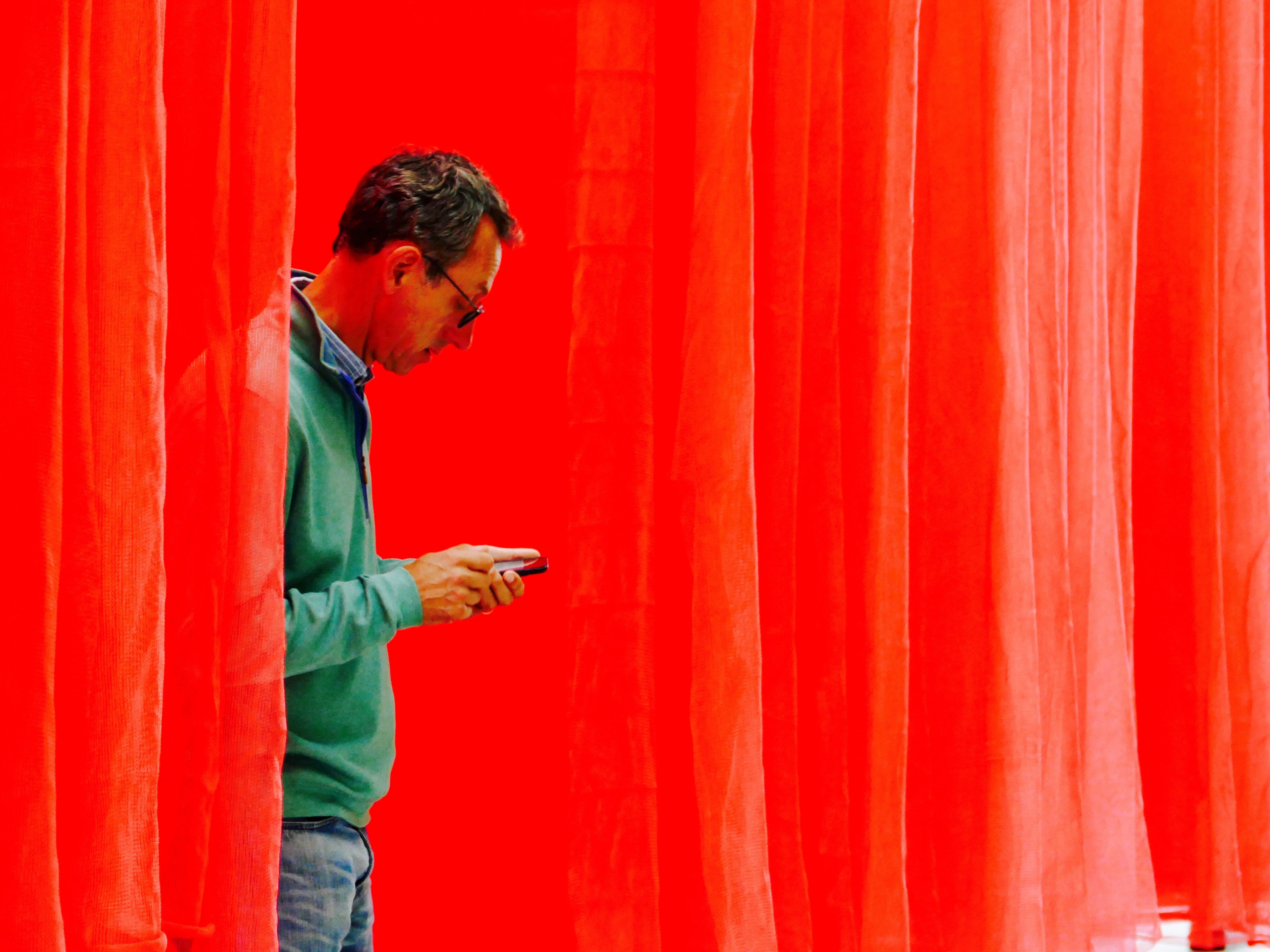 A man engrossed with his smartphone behind bright red curtains, lost in thought.