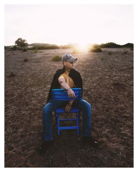 A man in a cap sitting on a blue chair outdoors during sunset, exuding a sense of solitude and contemplation.