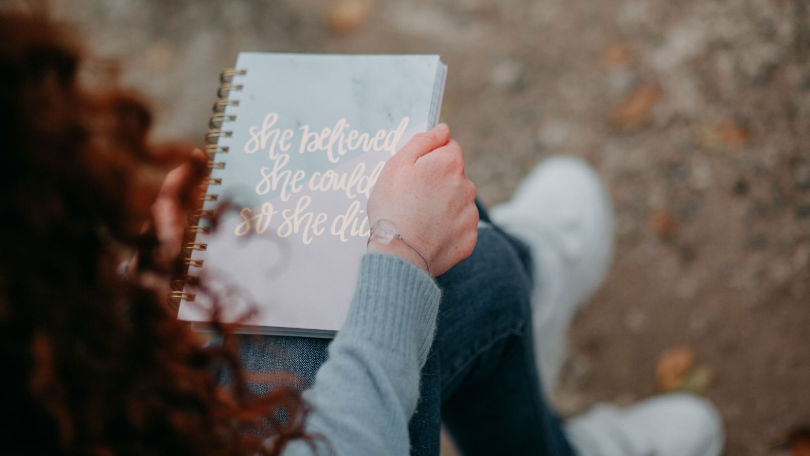 A woman sitting outdoors holding a notebook displaying an inspirational quote