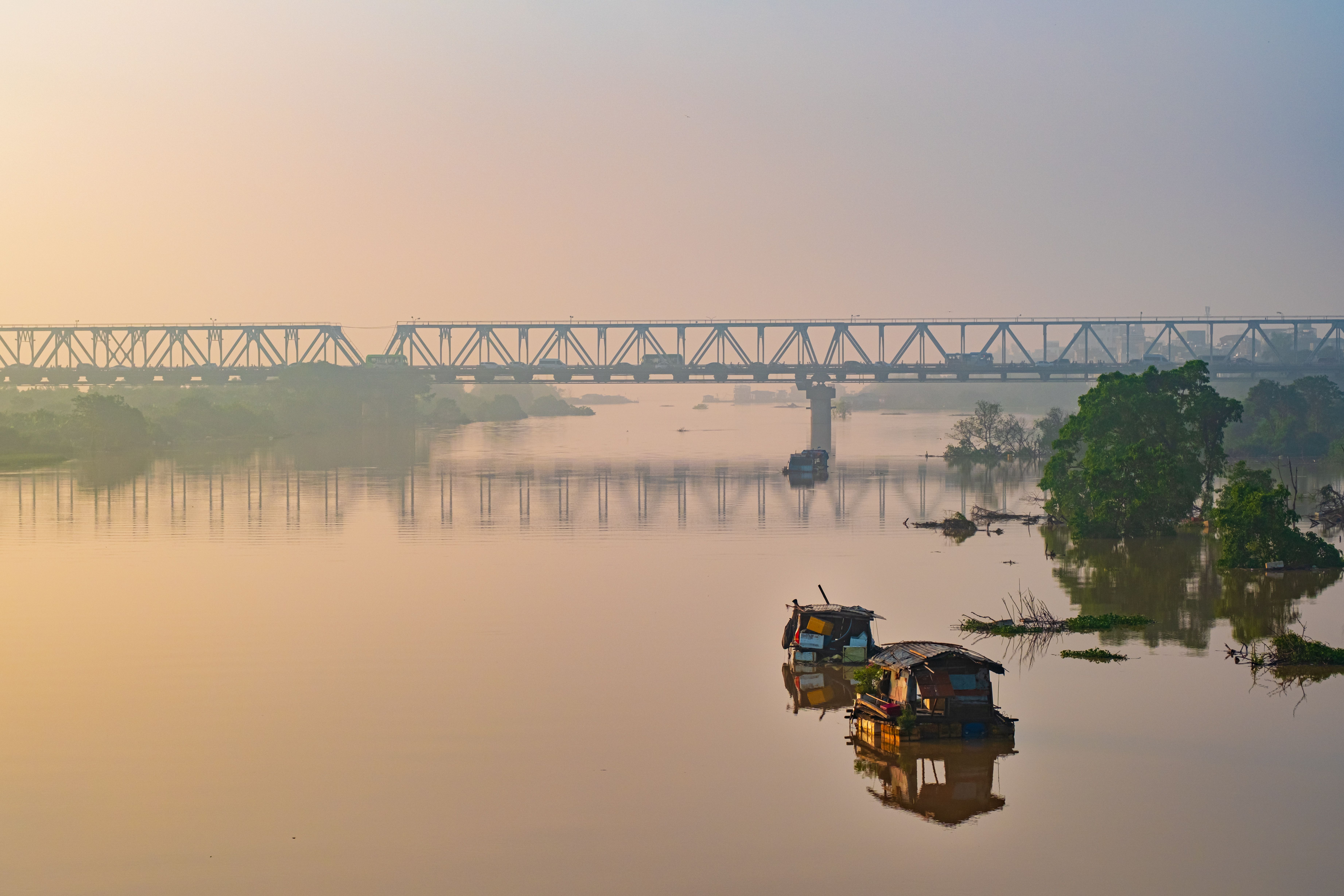 A tranquil river scene featuring a railway bridge, boats, and sunrise reflections on calm water.