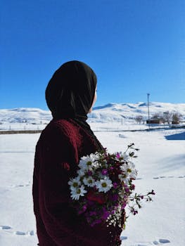 A woman in a hijab holds flowers against a snowy winter landscape under a clear blue sky.