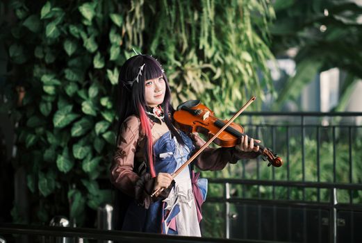 A cosplayer dressed in vibrant attire plays the violin amidst lush greenery, showcasing artistic expression.