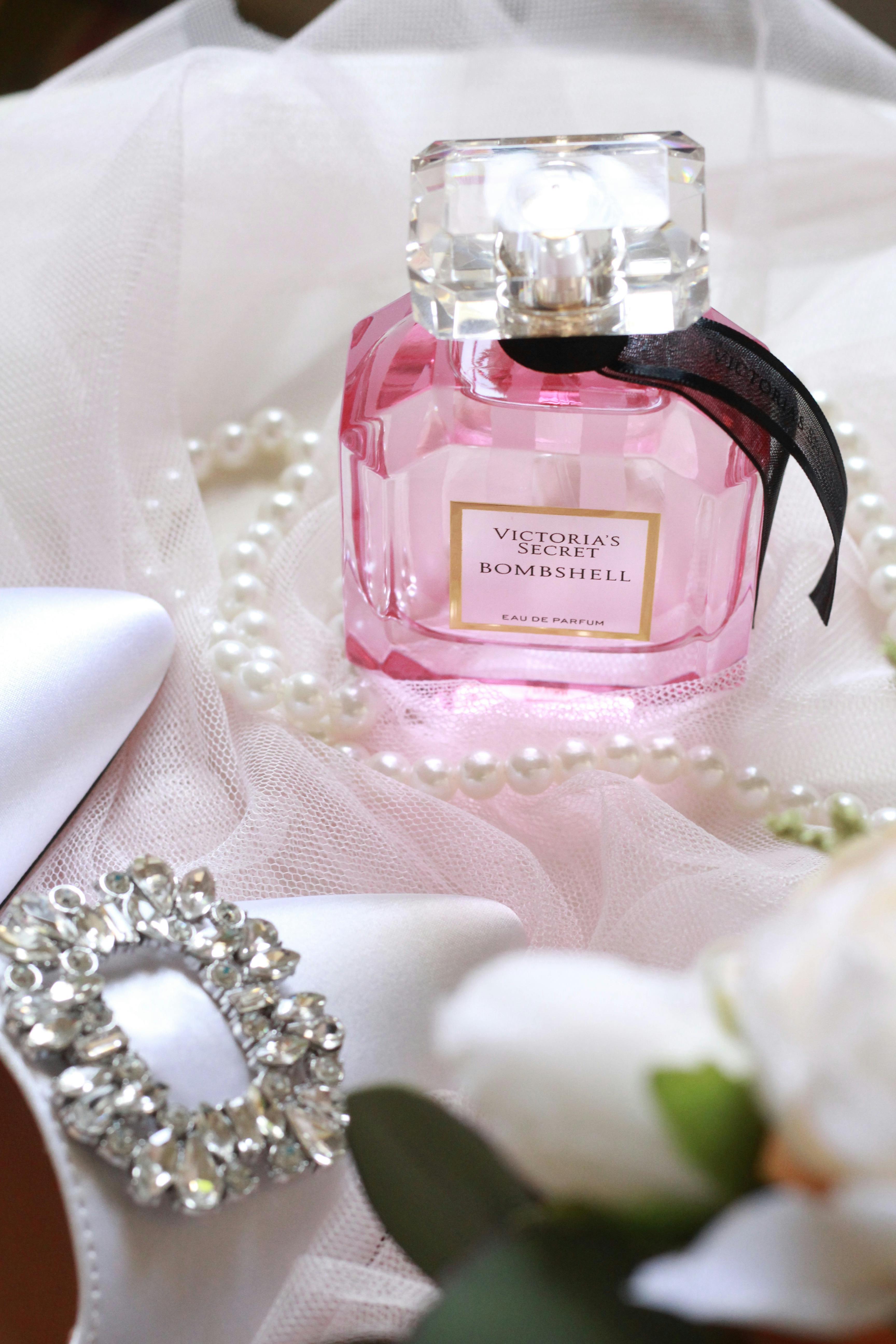A pink glass perfume bottle surrounded by pearls and white flowers on a soft fabric.