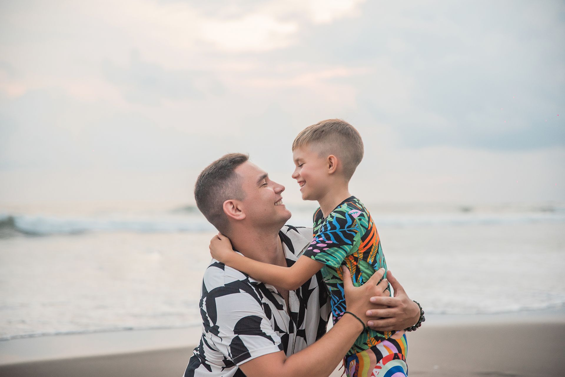 Father and son smiling at each other on the beach during a picturesque sunset, portraying family bonding and joy.
