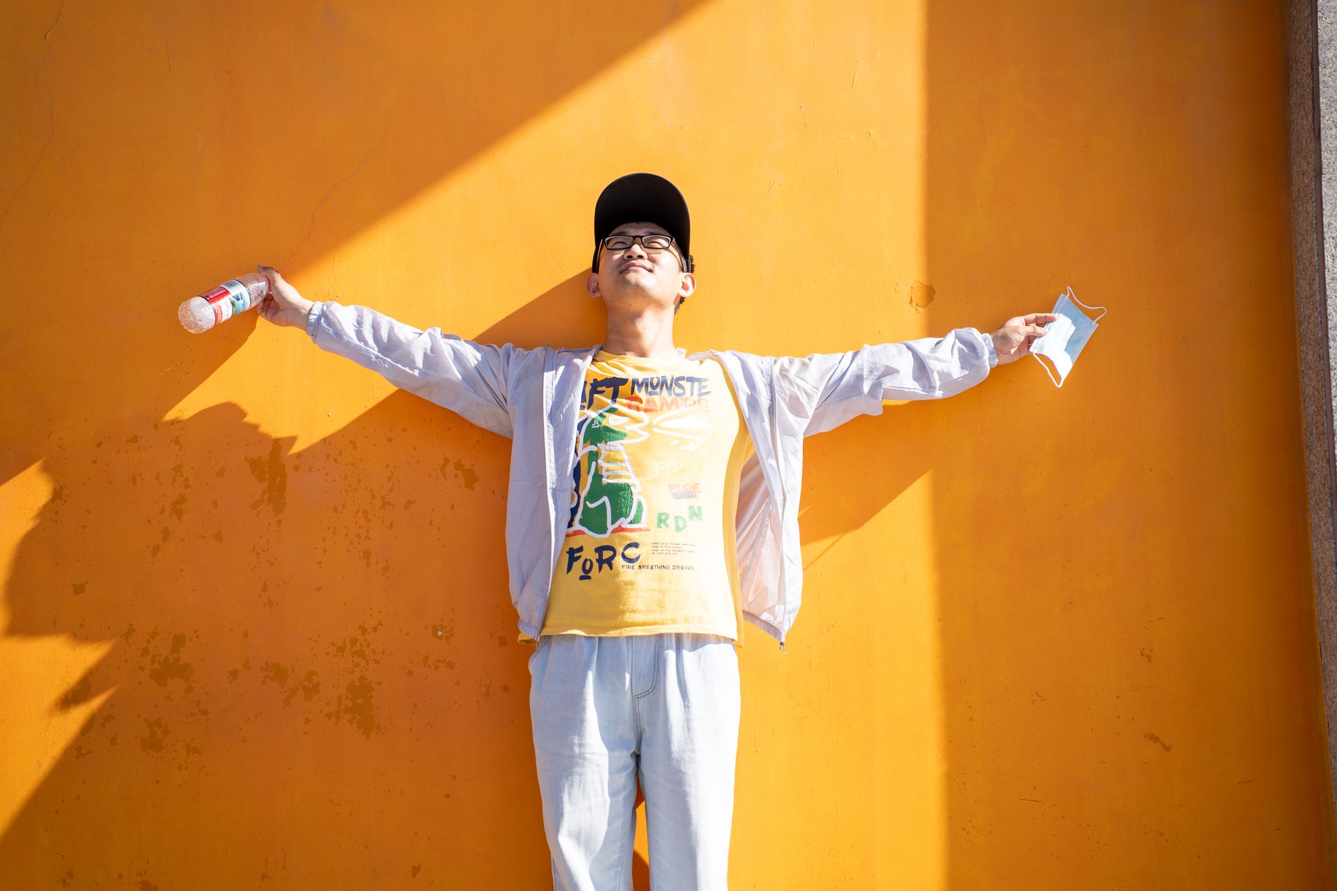 Young man in casual attire embraces sunlight against a vivid orange wall, exuding freedom.