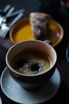 A close-up of a steaming cup of coffee paired with a slice of cake on a moody setting.