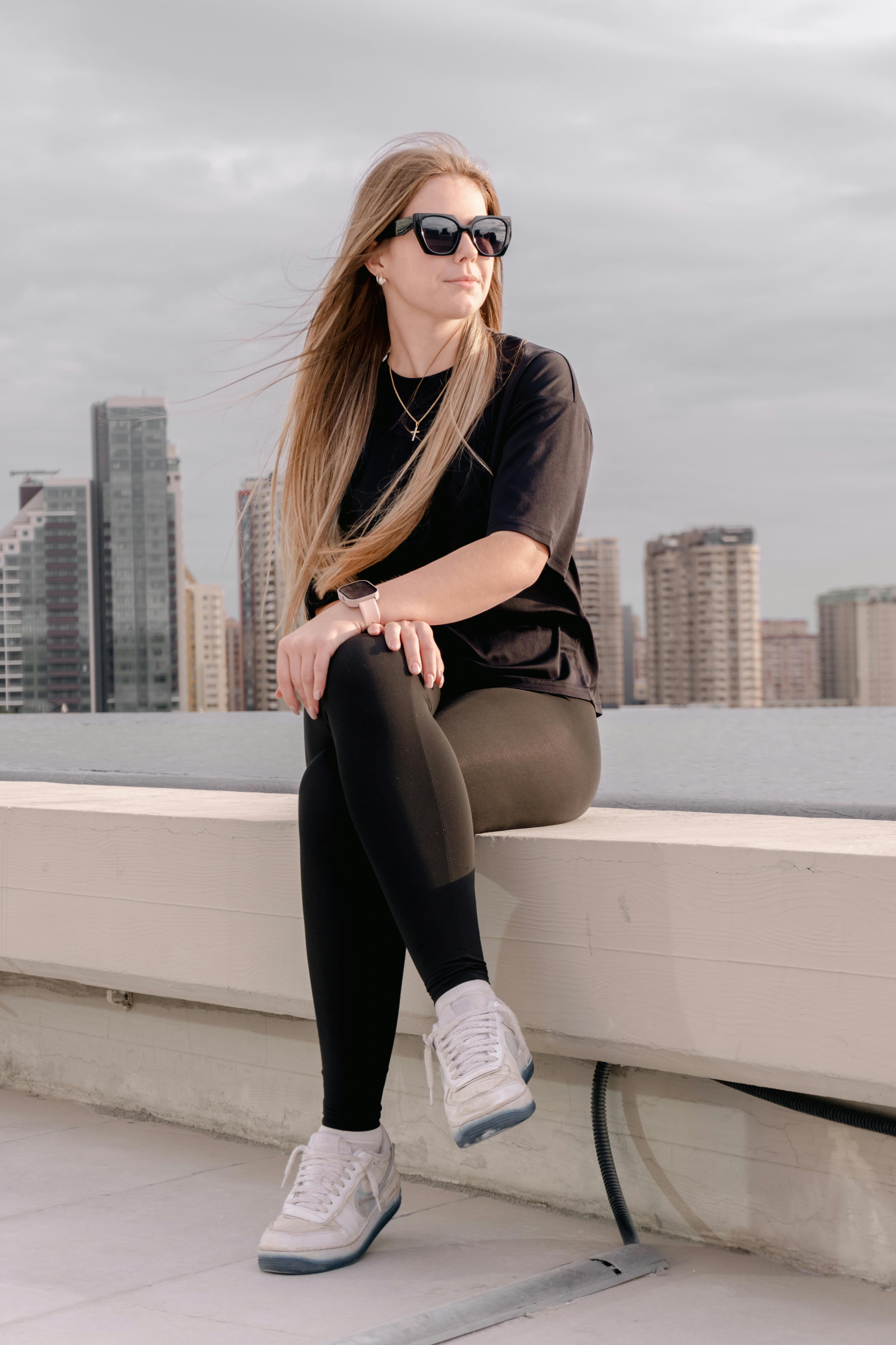 Woman sitting on a rooftop ledge with a city skyline view, wearing casual attire and sunglasses.