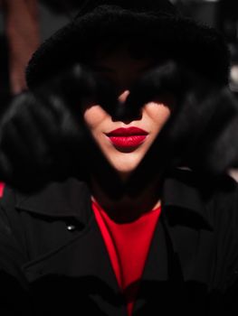 A mysterious woman with bright red lipstick forms a heart shape with black gloves.