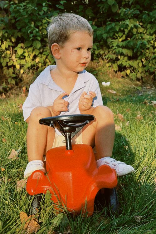 Free Cute young boy sitting on a red toy car in a sunny garden. Stock Photo