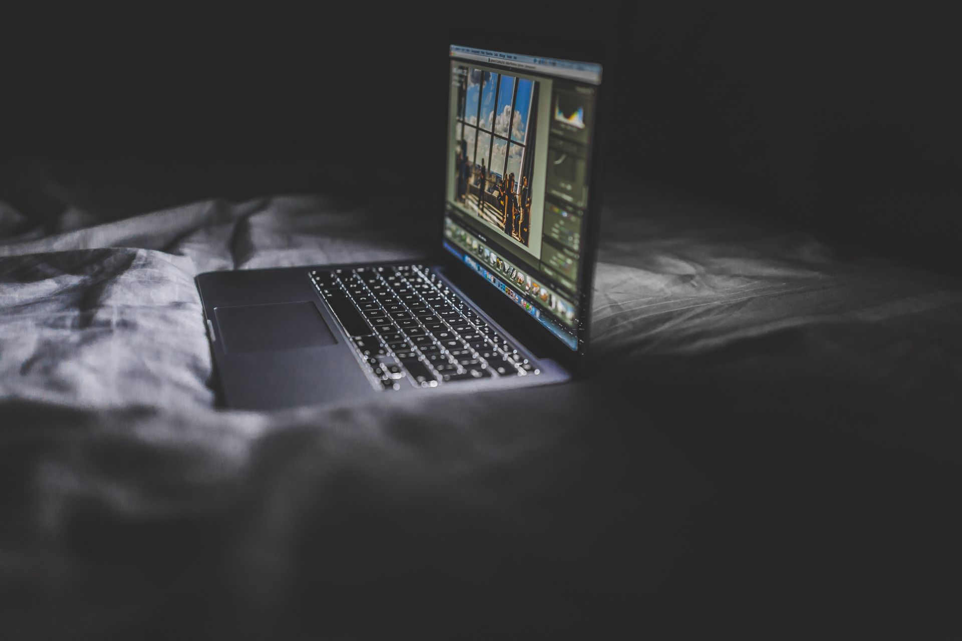 A laptop glowing on a bed at night with a city visible through the window.