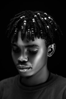 Striking black and white portrait of a young man with beaded hair set against a dark background.