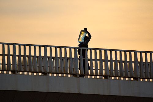Free A striking silhouette of a person carrying a water container across a bridge at sunset in Kampala, Uganda. Stock Photo