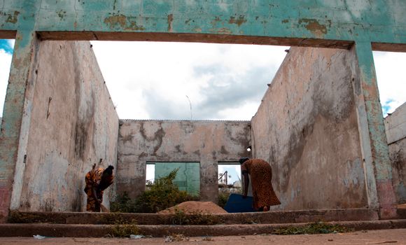 Two women cleaning rubble in a deserted, open-roof structure under cloudy skies.