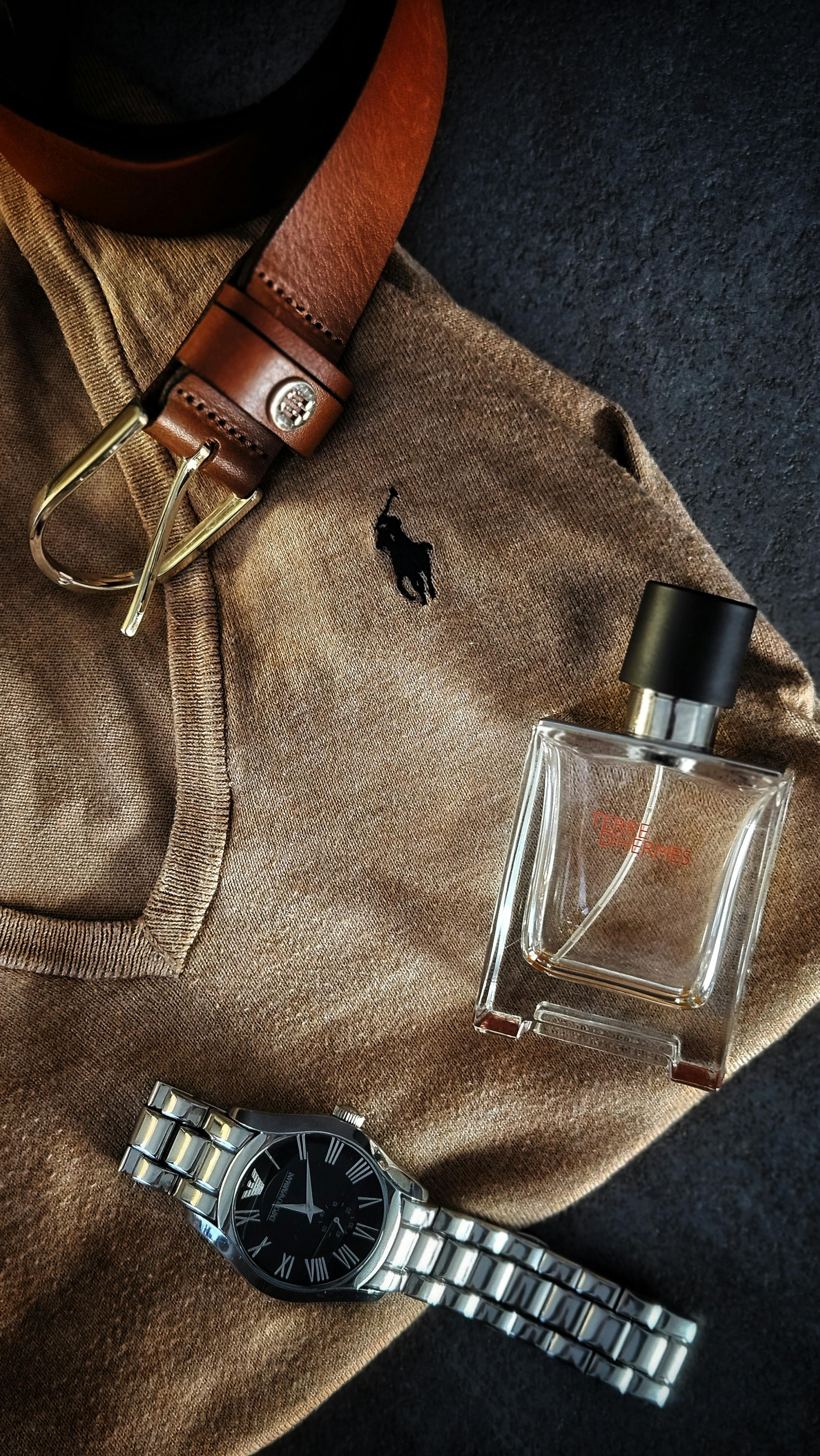 Flat lay of men's accessories including a brown sweater, leather belt, watch, and cologne for a classic style.