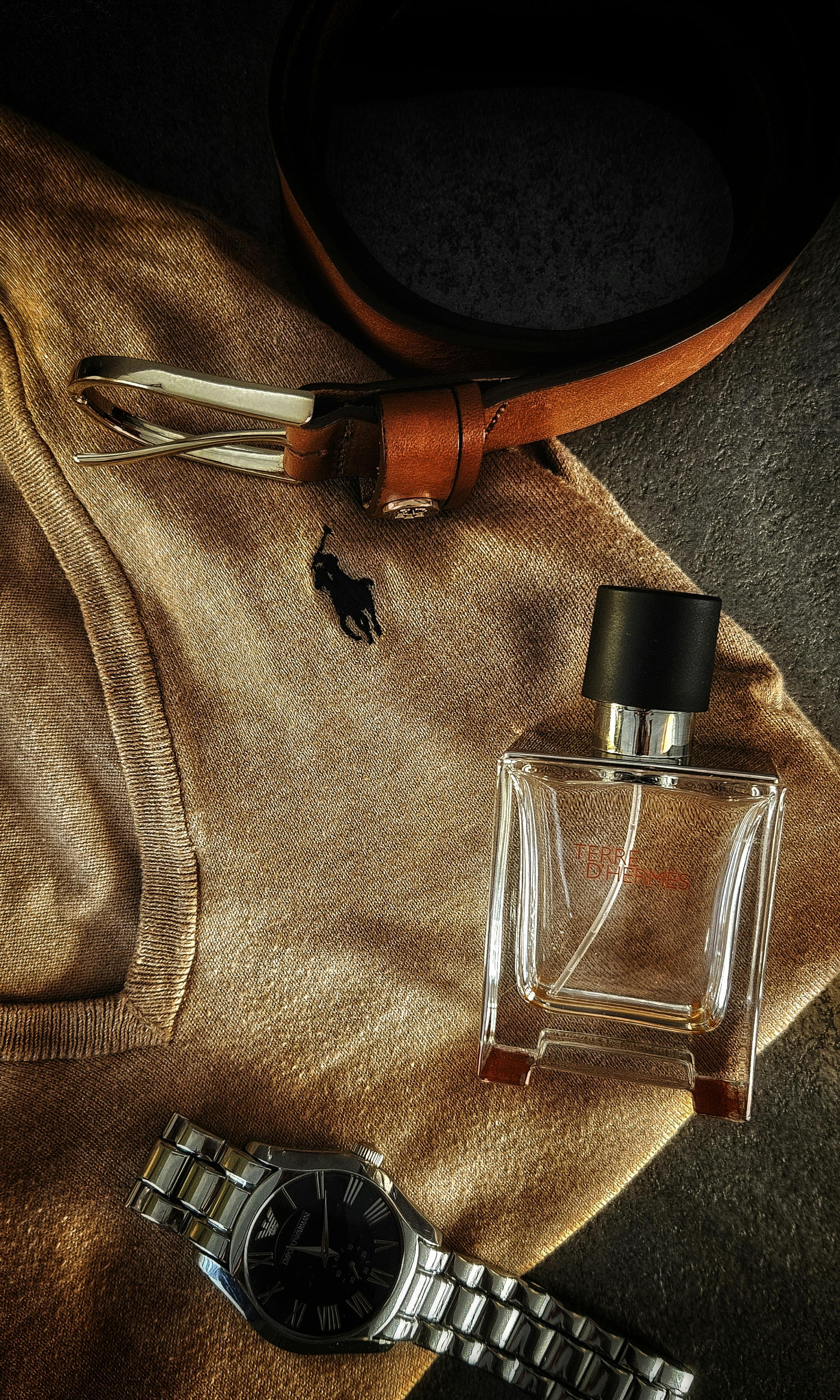 A stylish flat lay of men's fashion accessories featuring a belt, sweater, watch, and fragrance bottle.