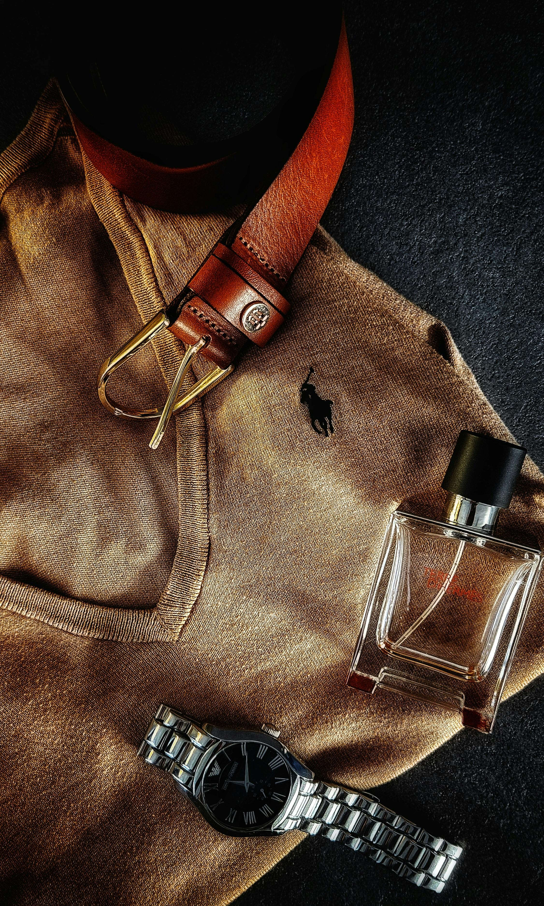 Stylish flat lay featuring a brown sweater, leather belt, wristwatch, and fragrance bottle.