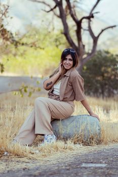A stylish woman sits on a rock in an outdoor setting in Tehran, Iran, enjoying a sunny day.