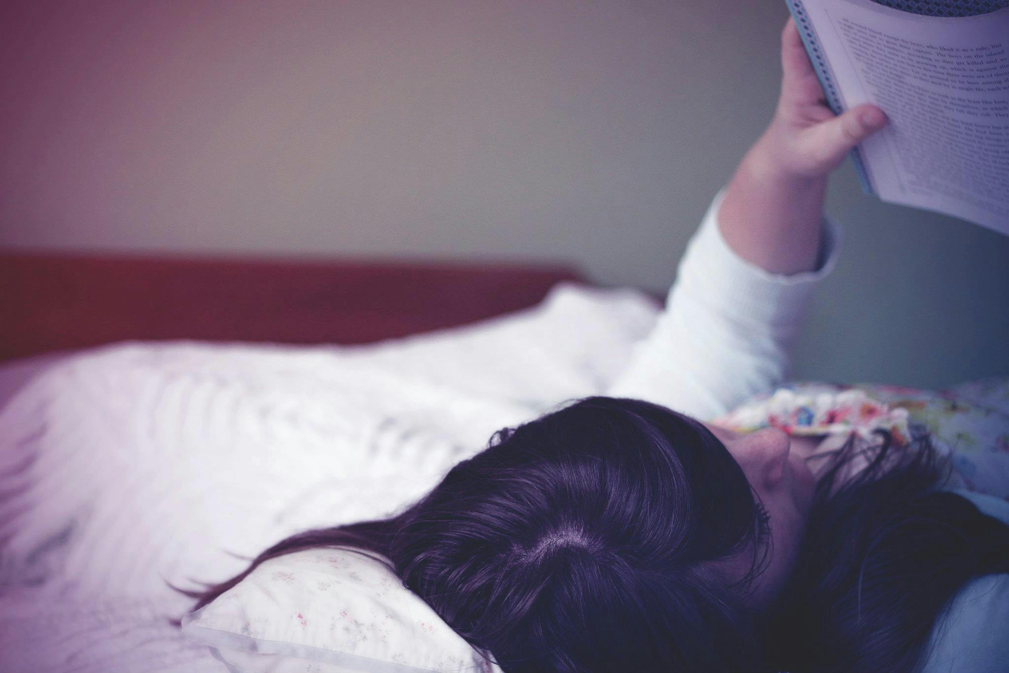 Woman Holding Book While Lying on White Bed Sheet