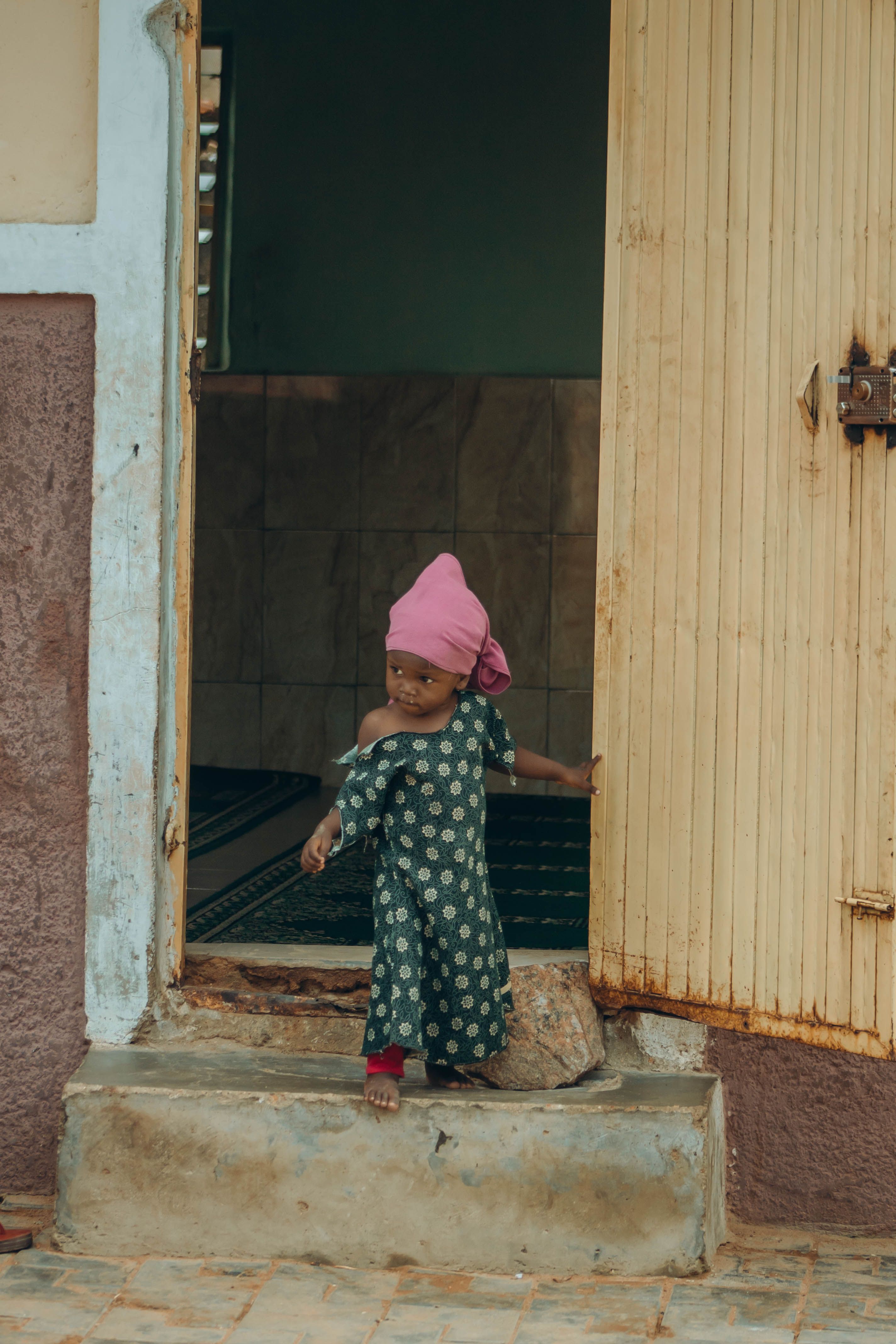 A young child in a patterned dress and headscarf stands in a doorway with a curious expression.