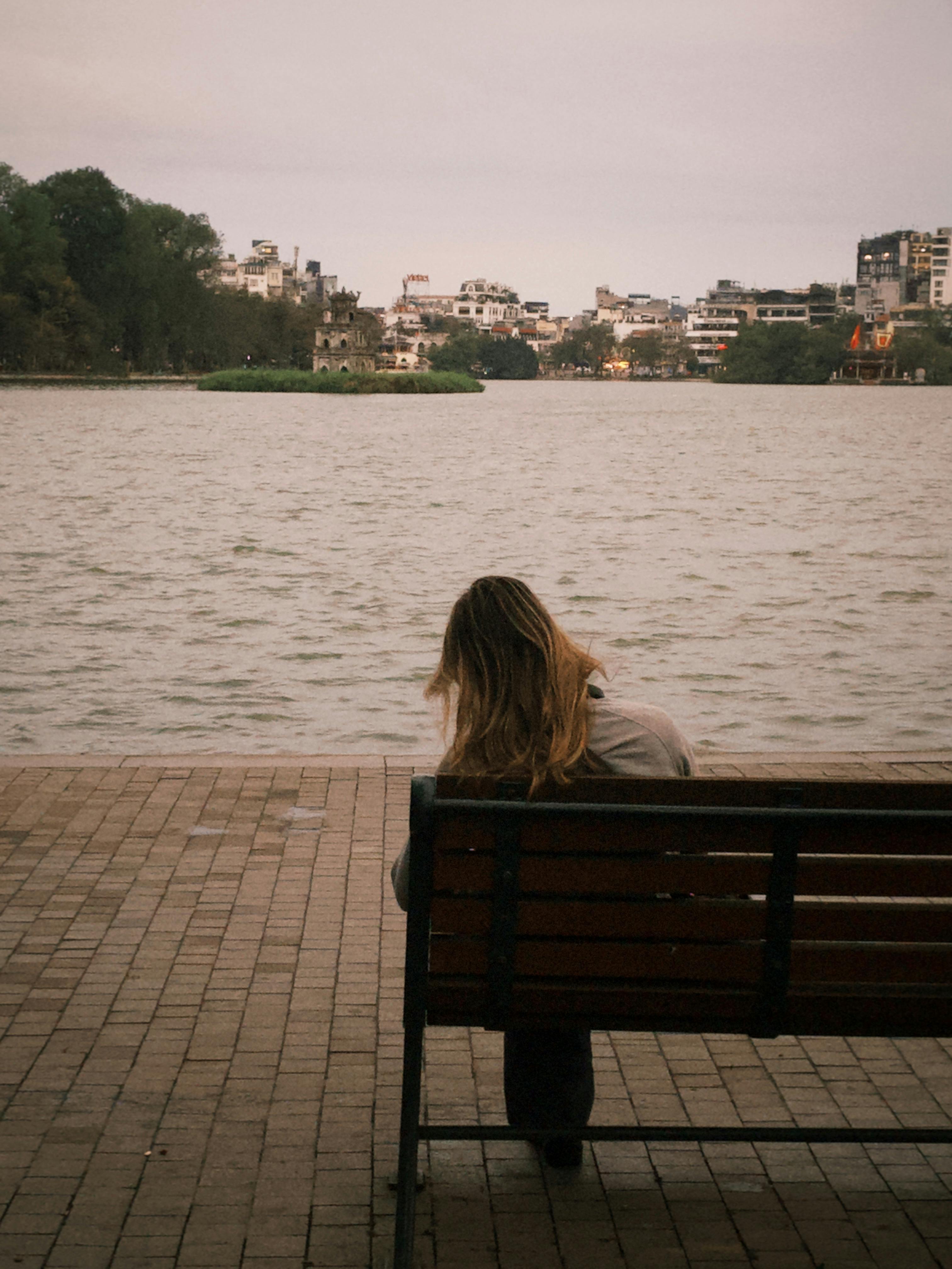 Woman sitting on bench by lake, urban skyline in background, peaceful solitude.