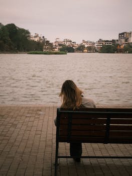 Woman sitting on bench by lake, urban skyline in background, peaceful solitude.