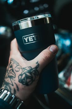 Close-up of a tattooed hand gripping a YETI tumbler in an outdoor setting.