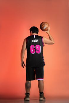 A man in a black and pink jersey holding a basketball against an orange background.
