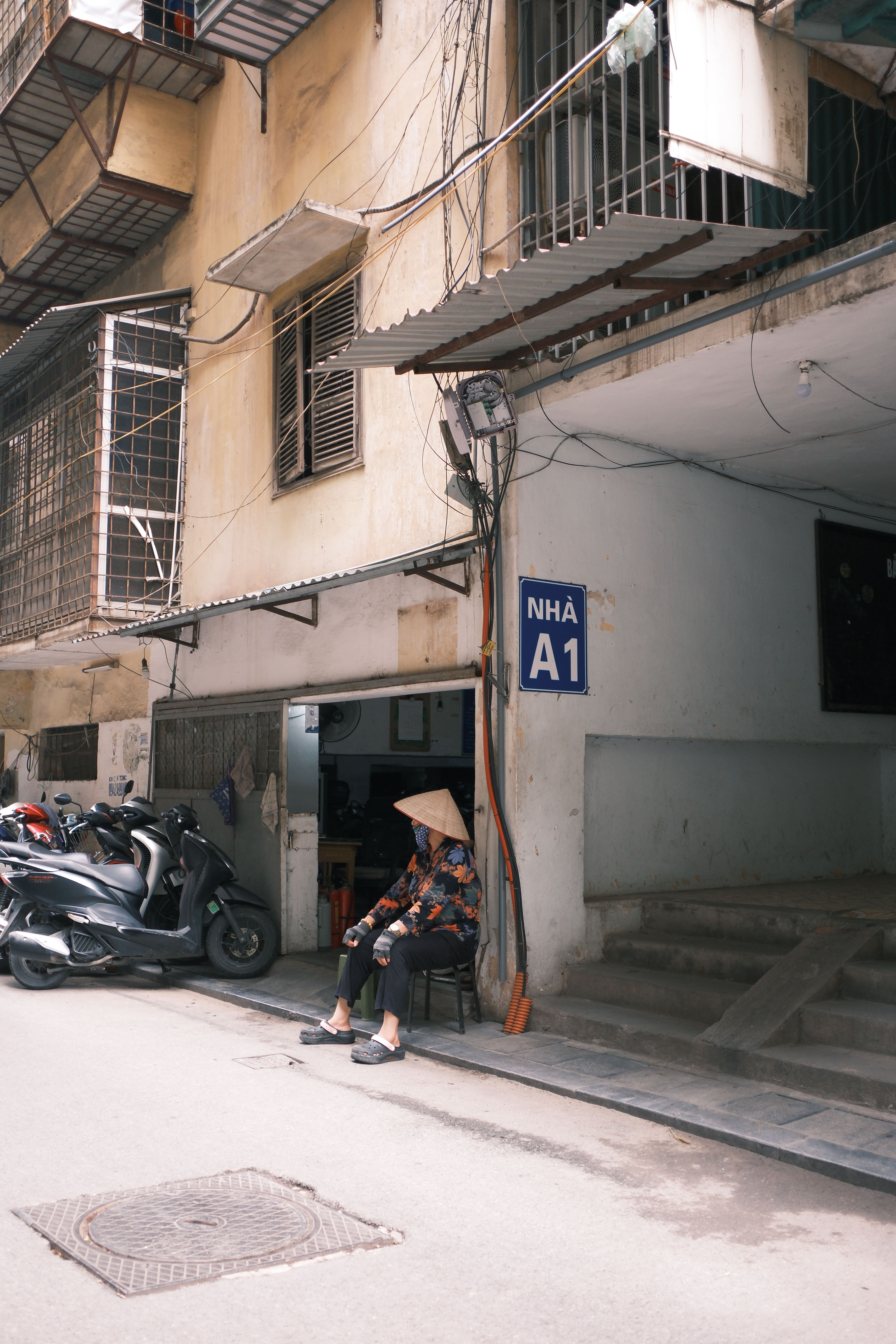 An elderly woman in a conical hat sits in an urban street in front of a building in Jaipur, India.