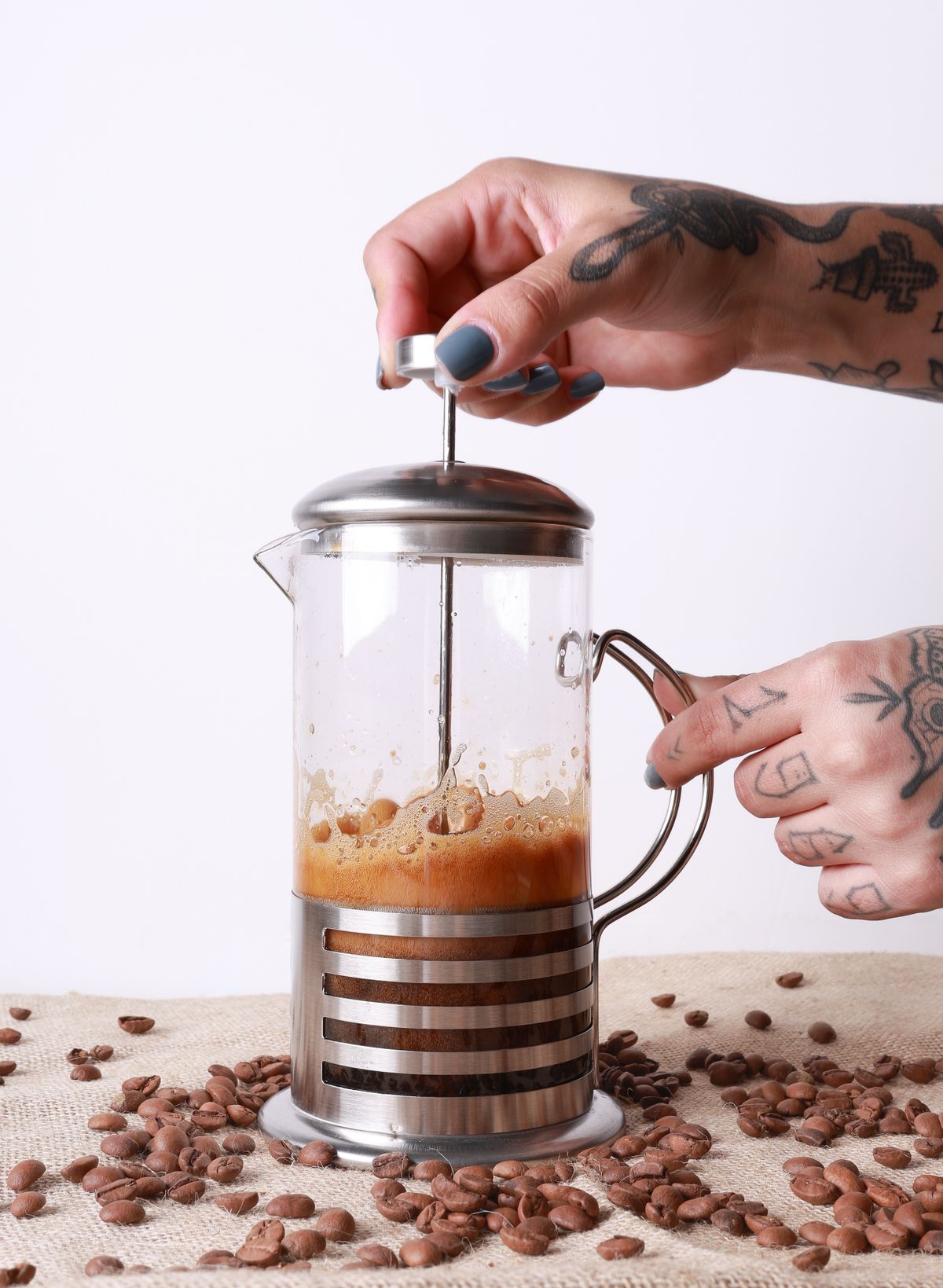 French Press din sticla cu cafea proaspat infuzata si boabe imprastiate pe masa