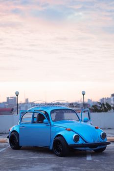 A classic blue car on a rooftop with open doors, set against a cityscape during sunset.