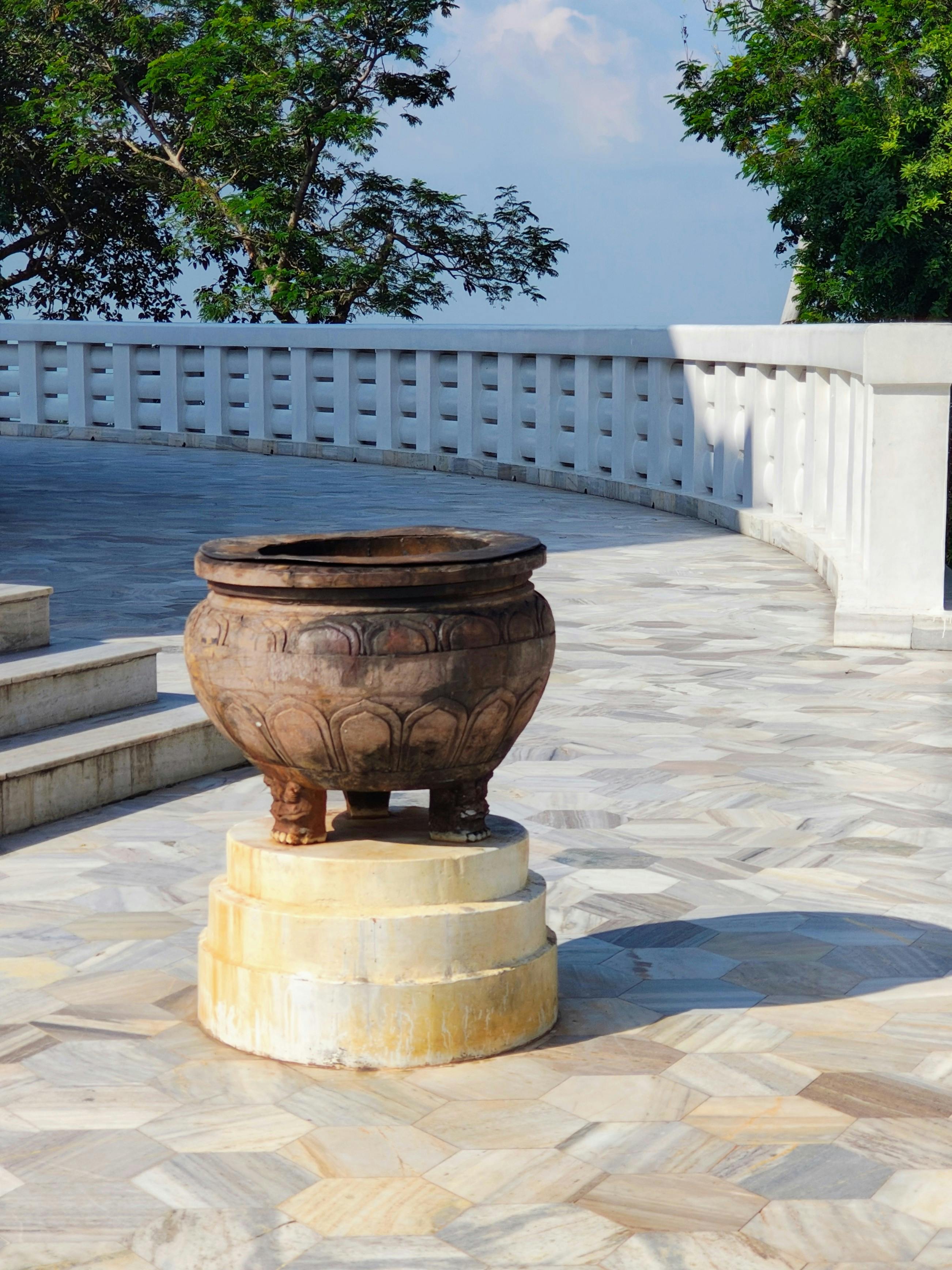 Antique metal pot on a tiered marble base on a sunny stone terrace with a white railing and trees in the background