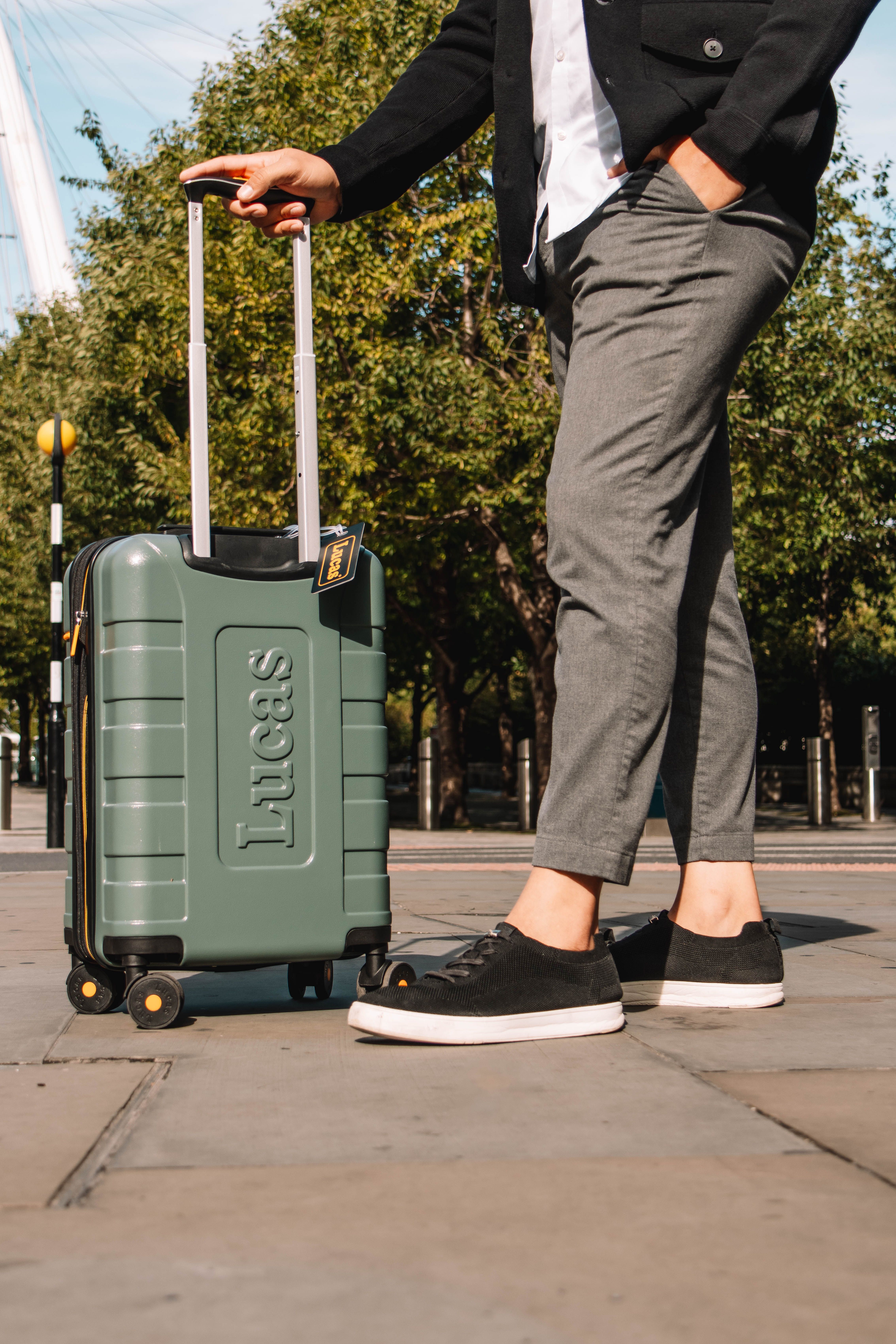 Person standing with suitcase on city sidewalk, trees in background.