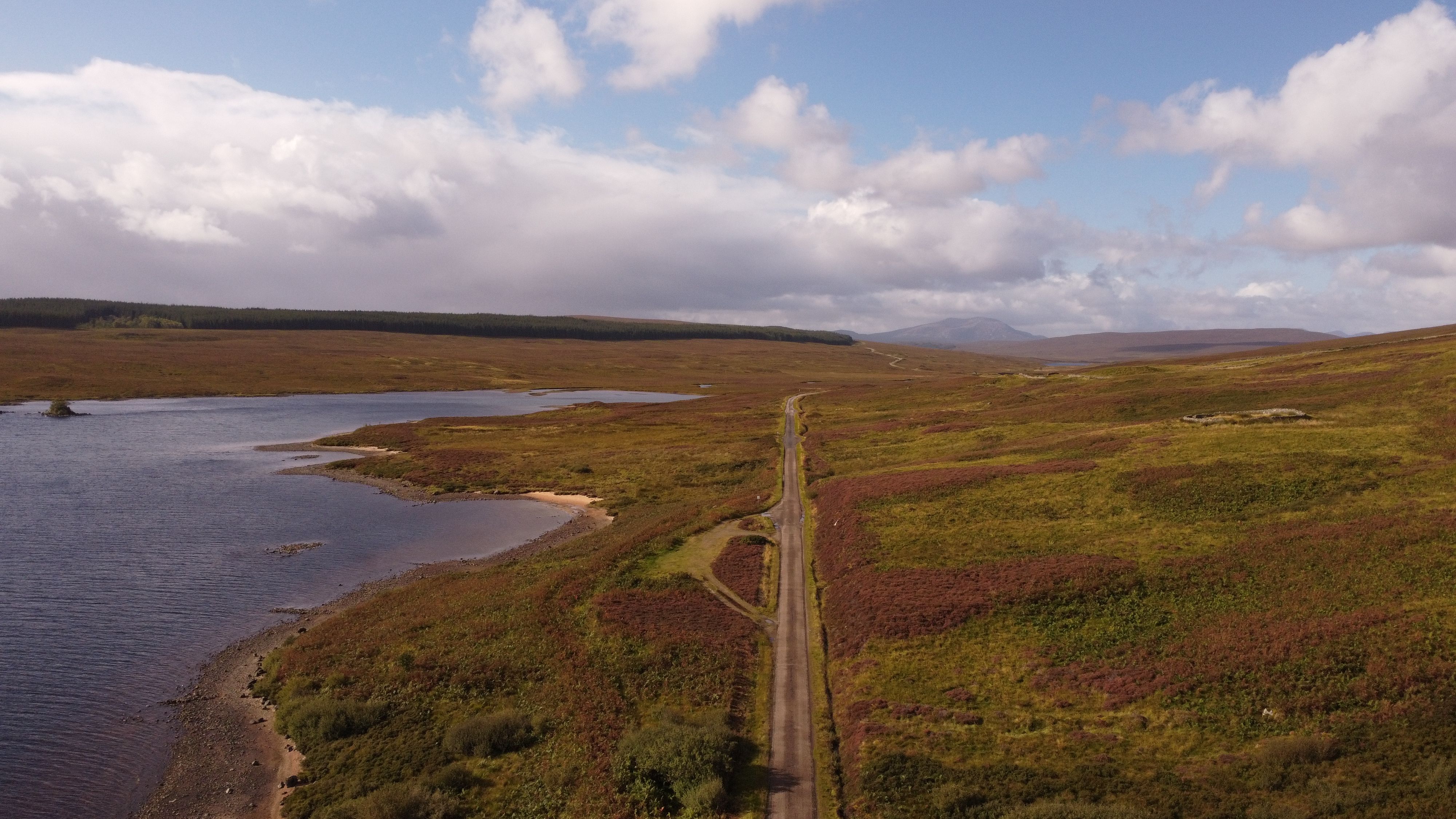 Aerial view of remote road by a loch in Scotland under a cloudy sky.