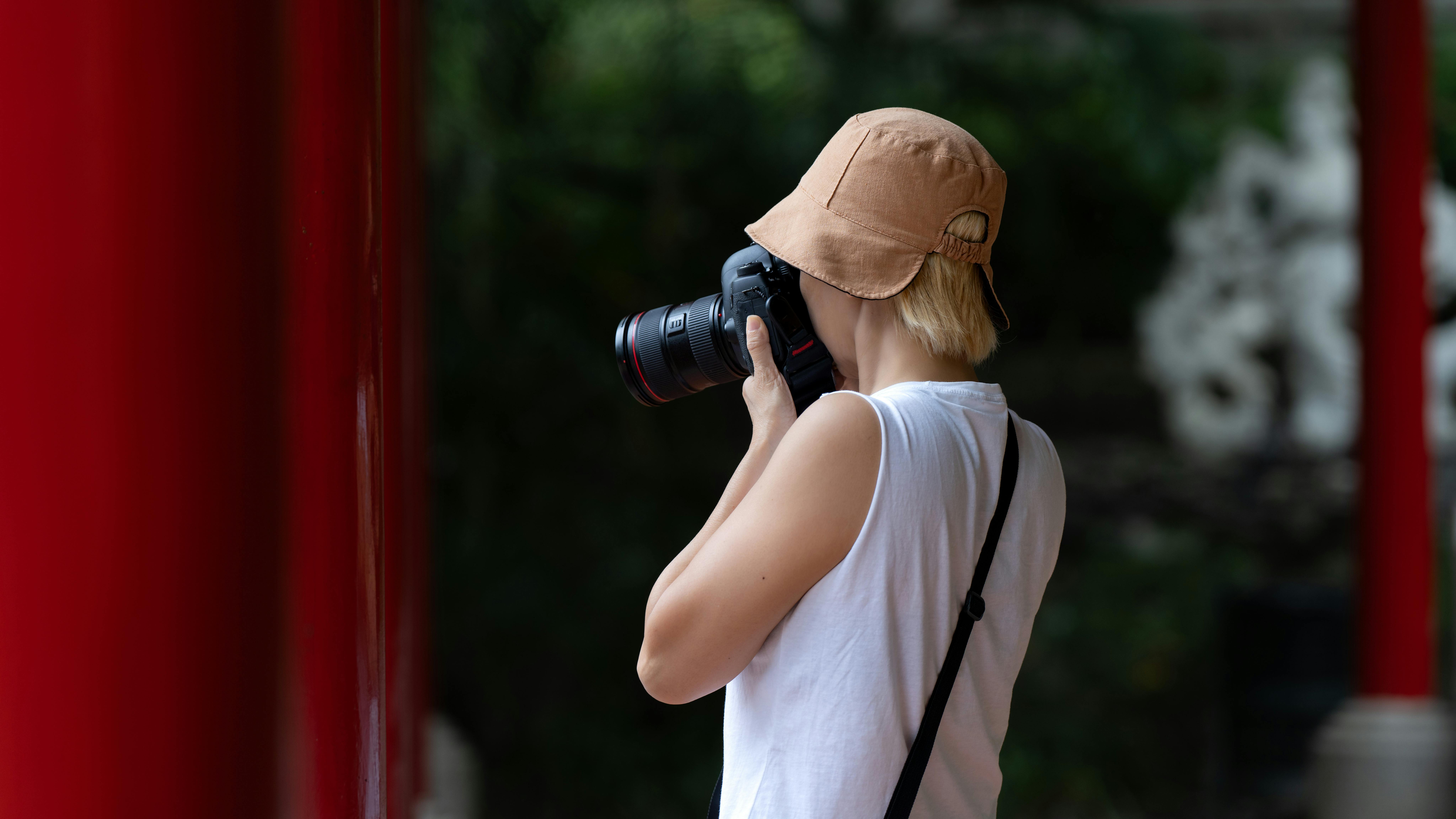 Free A photographer in a hat takes pictures outdoors with a DSLR camera, from a behind angle. Stock Photo