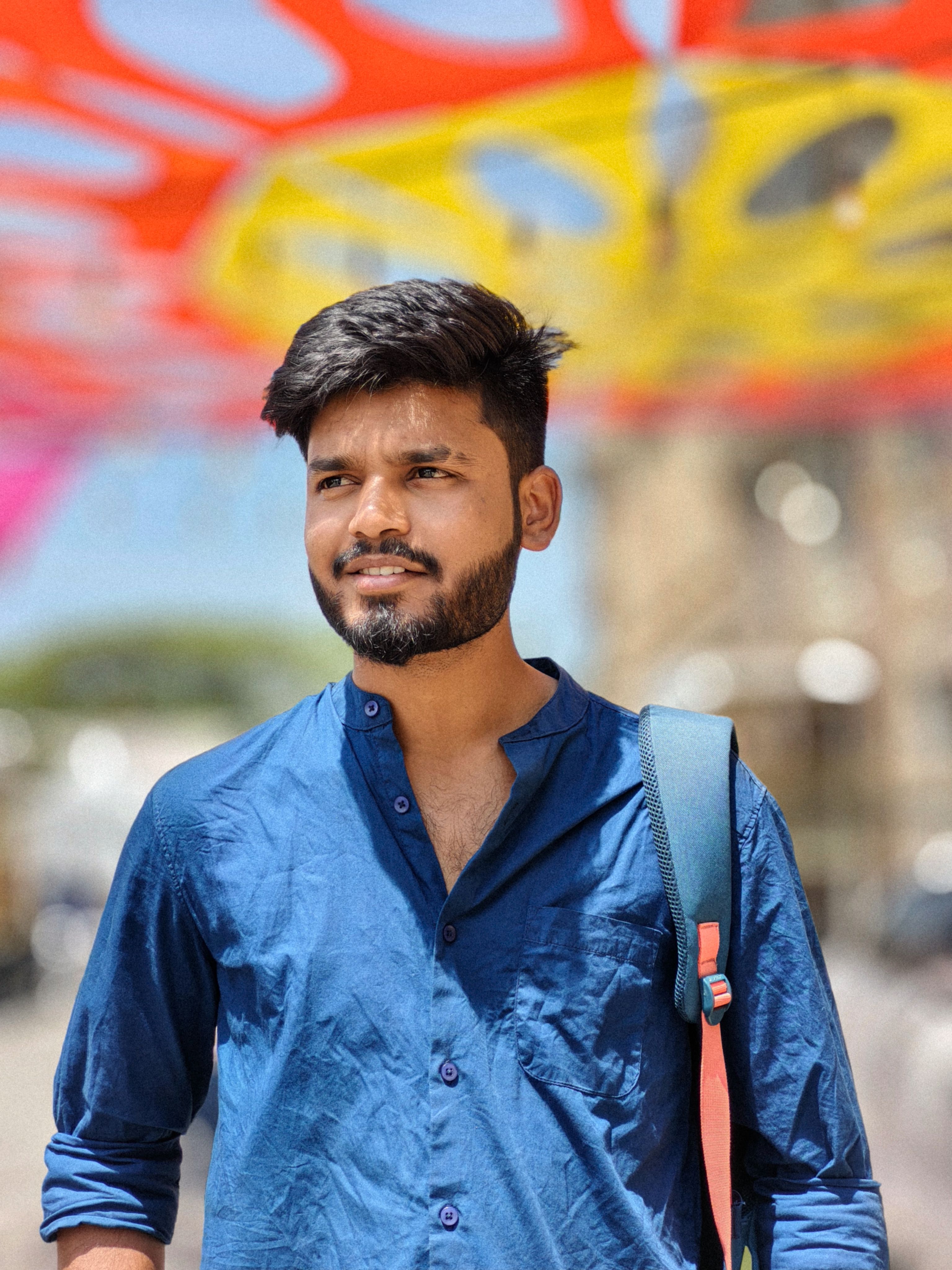 Portrait of a young man standing outdoors under a vibrant, colorful canopy on a sunny day.