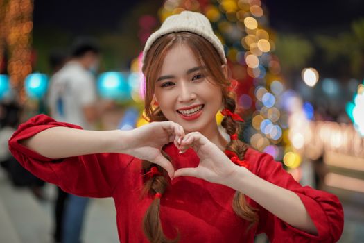 A joyful woman in festive attire making a heart shape with hands in a vibrant Christmas setting.