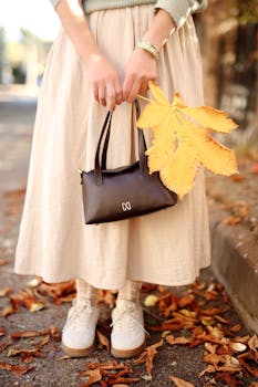 Stylish autumn outfit with a yellow leaf and handbag on a leafy street.