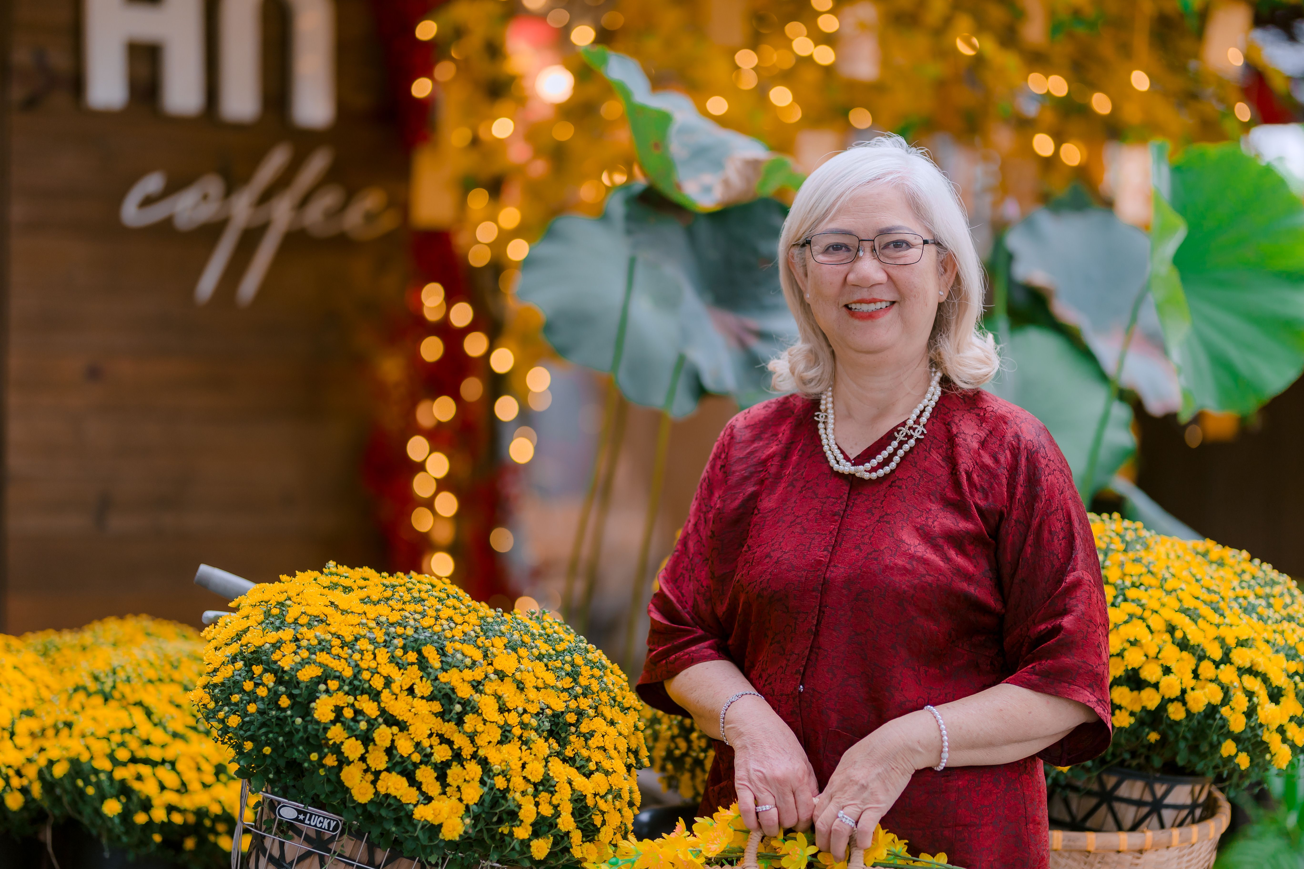 A senior woman smiling with vibrant yellow flowers in a festive outdoor setting.