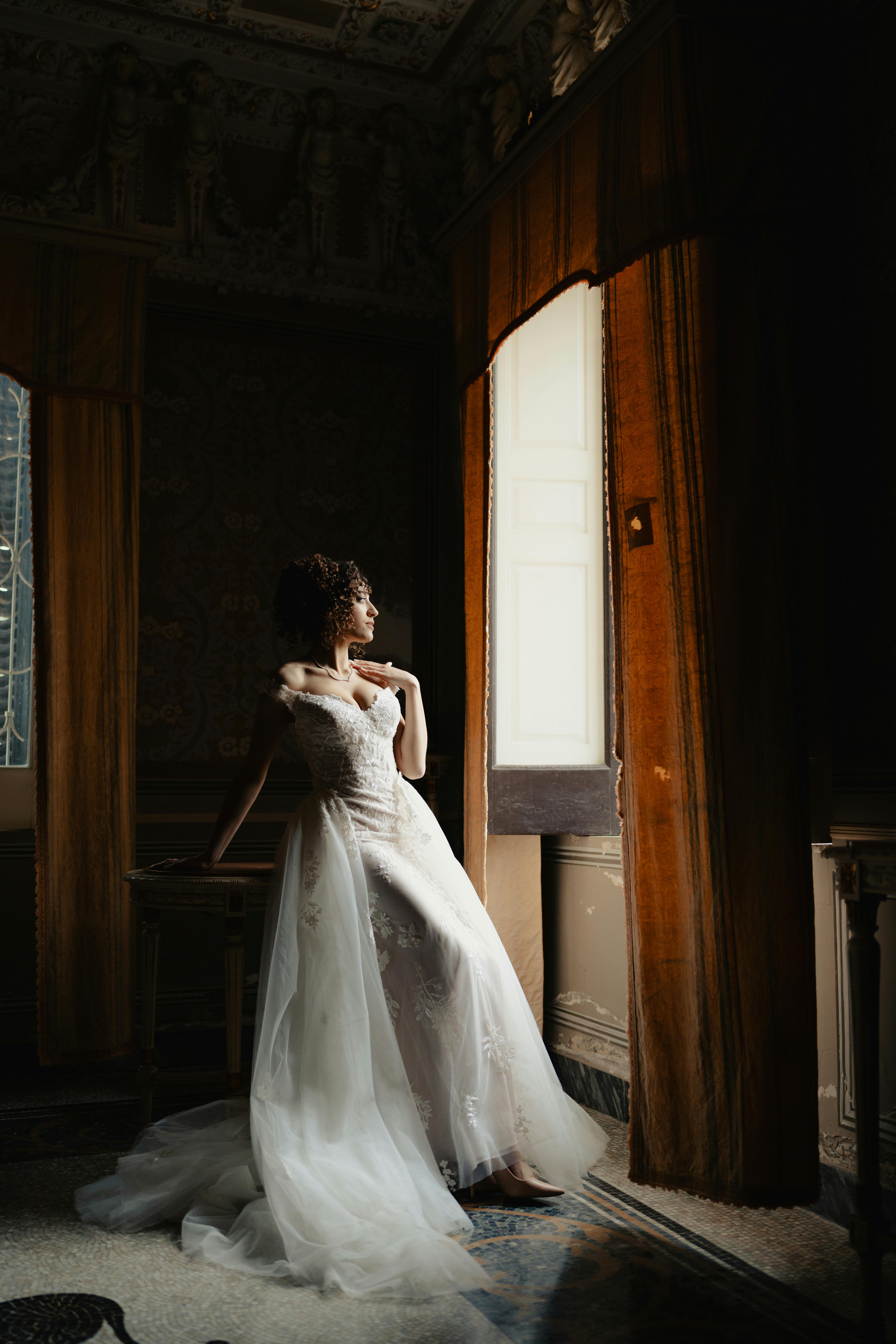 A bride in a vintage room gazing out of a window with soft natural light.