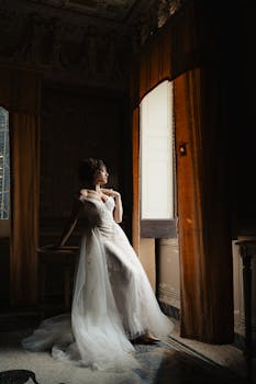 A bride in a vintage room gazing out of a window with soft natural light.