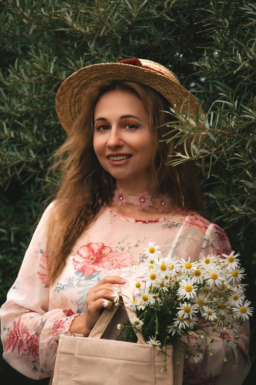 Free Smiling woman in floral dress and straw hat holding daisies in a garden setting. Stock Photo