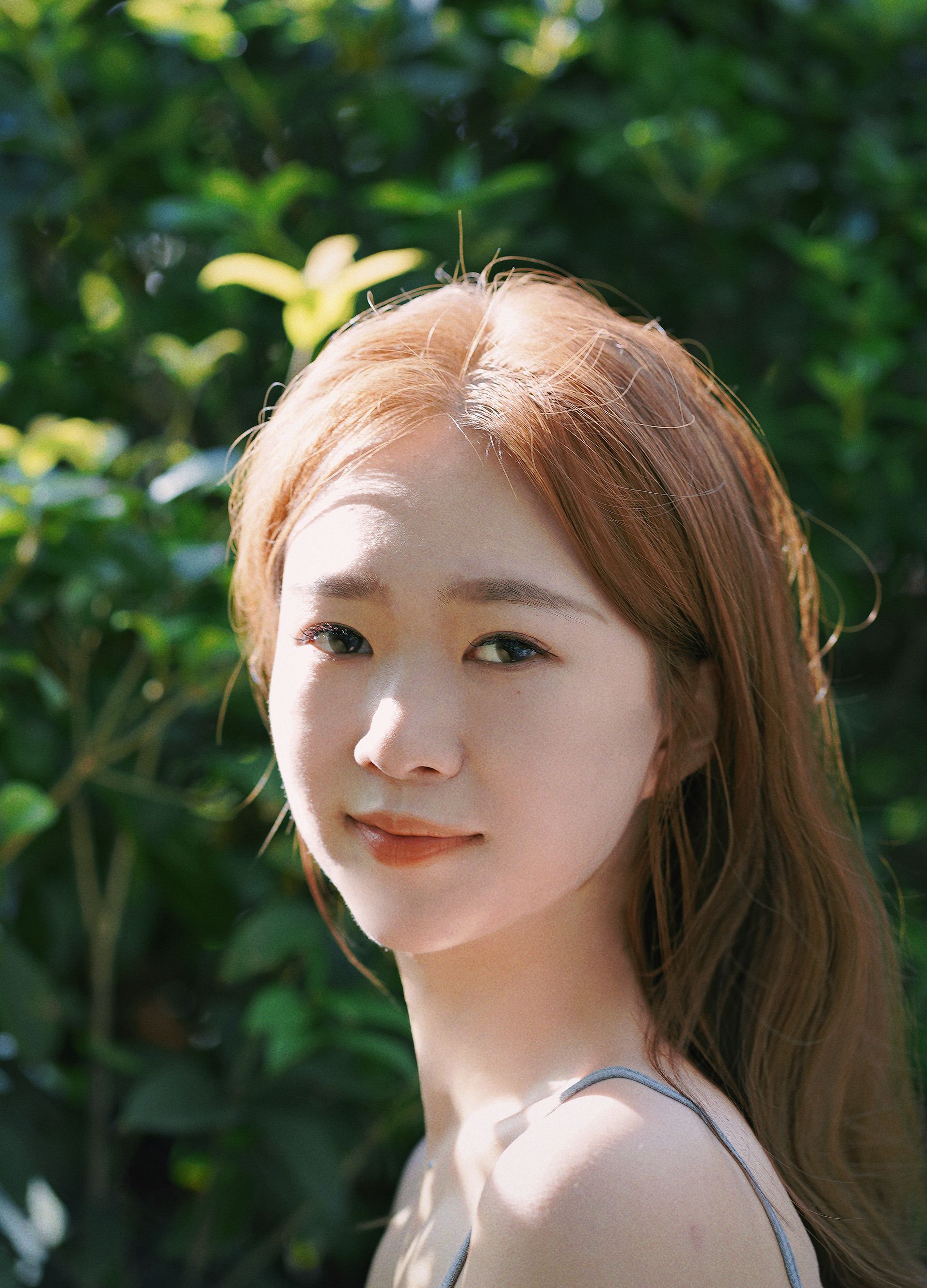 Portrait of a young woman smiling outdoors in natural sunlight with green leaves background.