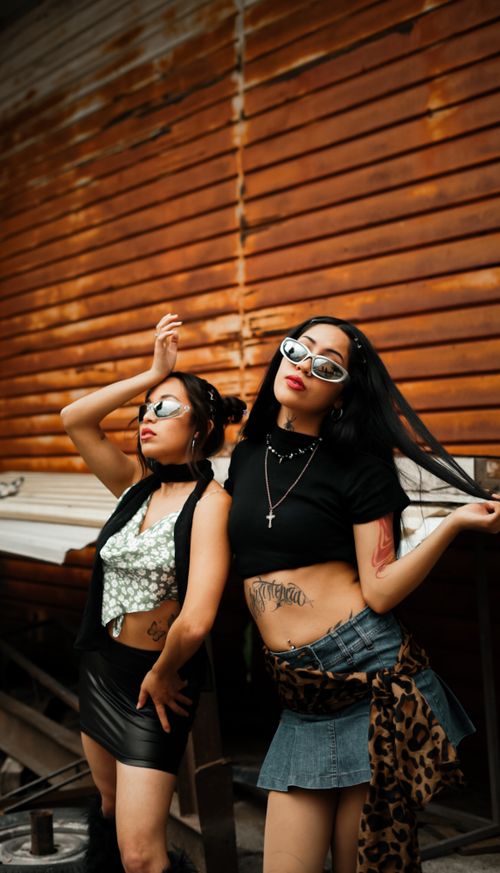 Free Fashion-forward women posing confidently outdoors against a wooden wall, showcasing tattoos and trendy outfits. Stock Photo